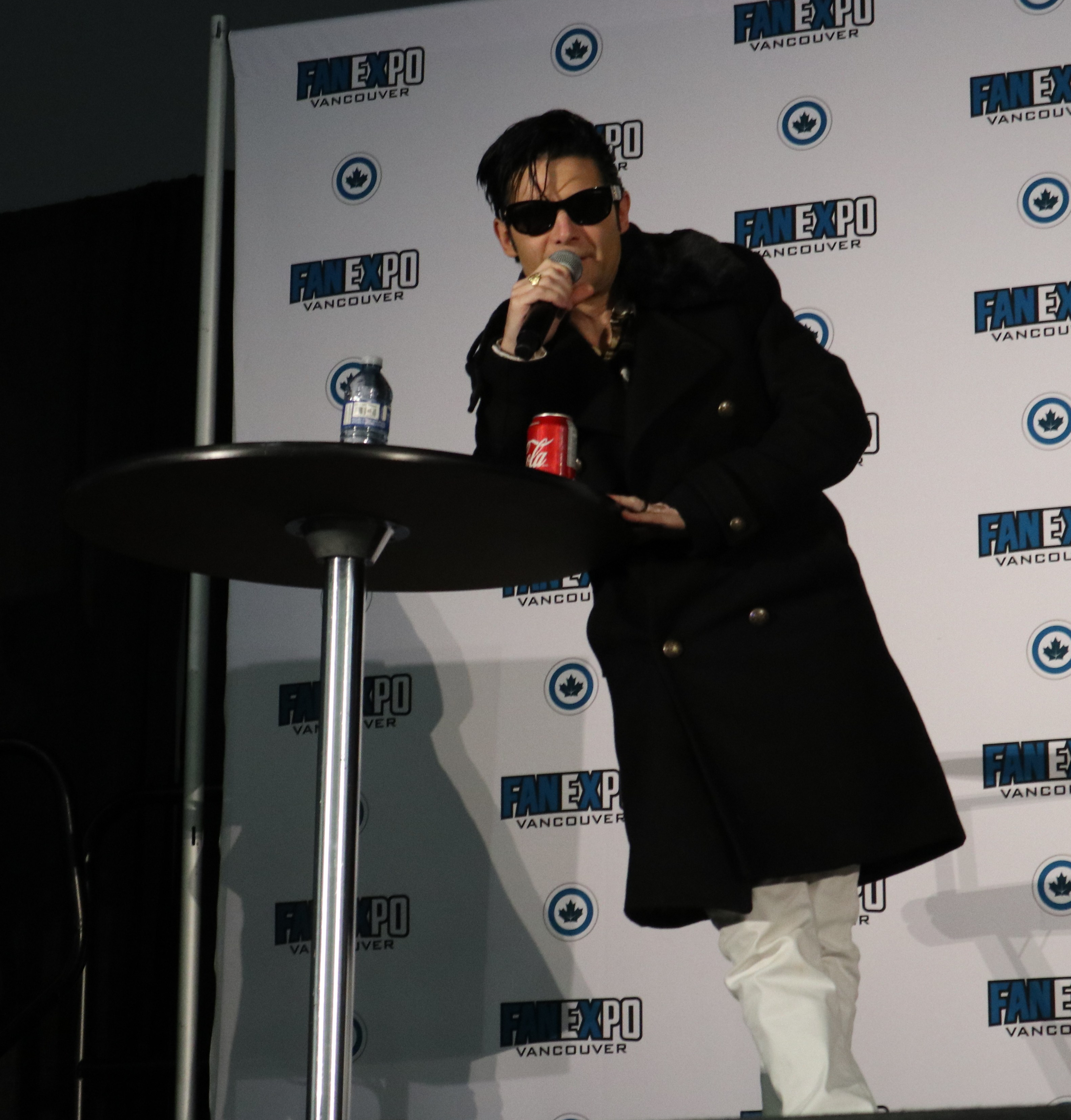 Corey Feldman leans against a high table. He is a Caucasian male with black hair that falls into his face. He is wearing dark sunglasses. He is wearing white pants and a long black coat. He is holding a microphone to his mouth to speak.